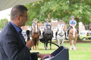 Governor Matt Meyer stands at a podium holding a microphone. Side view, the Governor is in the foreground and a line of police horses stands in the background.