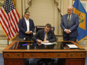 Governor Meyer sits at his desk signing legislation with House Majority Leader Kerri Evelyn Harris standing to the left of him and Secretary of Finance Michael Smith to the right.