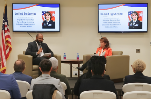 Richard M. Potter, Jr. (left) and Brigadier General (Ret.) Karen Berry (right) are seated on a small stage, holding microphones during the Veterans Day Fireside Chat. Two TVs behind them display a photo of Berry with the text ‘Unified by Service: A Virtual Fireside Chat’ and her title. The backs of the audience’s heads are visible.