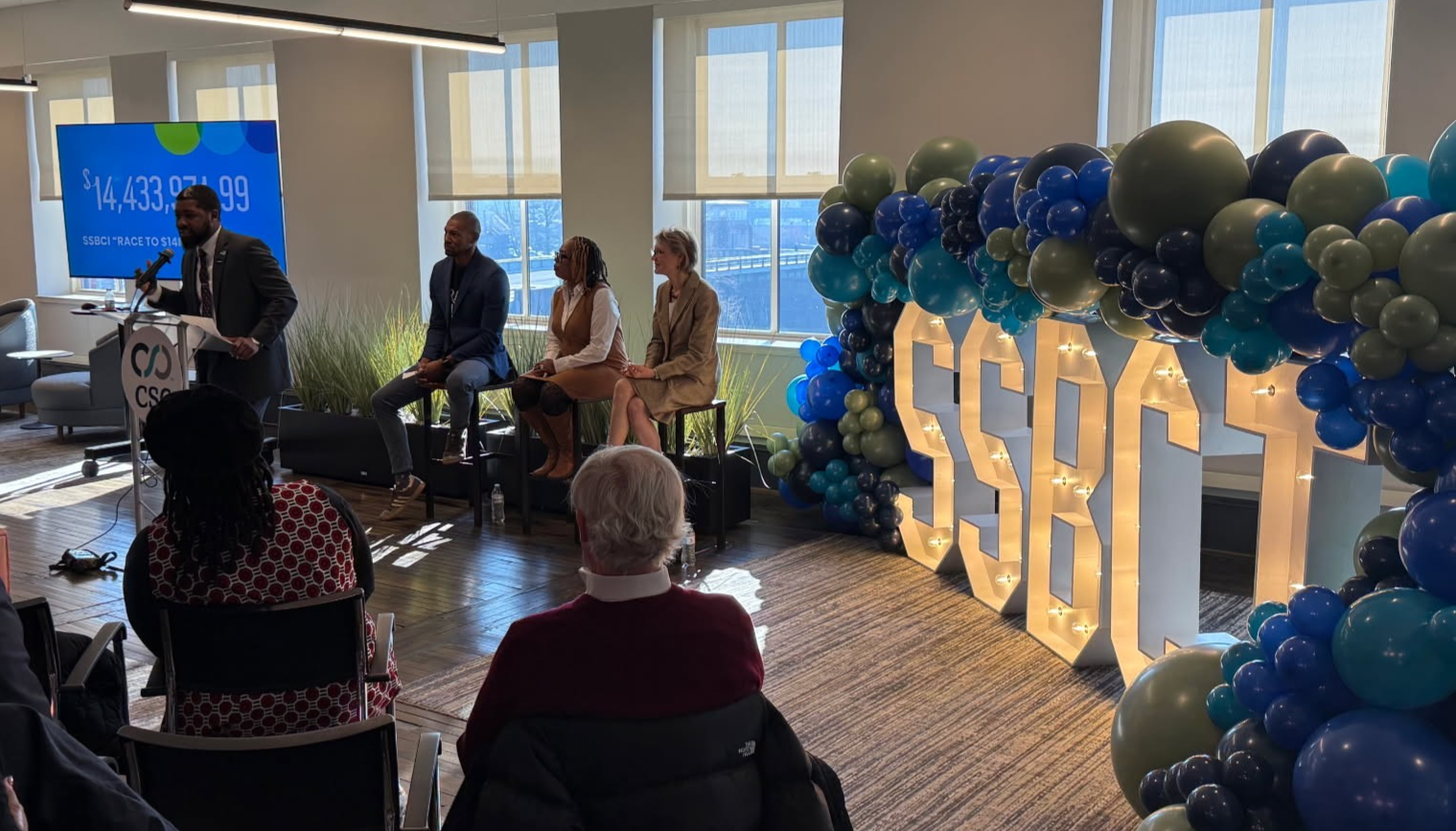 Director CJ Bell at the podium speaking to folks gathered to celebrate reaching our goal of $14M pushed out to small businesses in 8 months. Three speakers sit on stools beside him. A balloon arch sits on top of large lit up letters SSBCI.