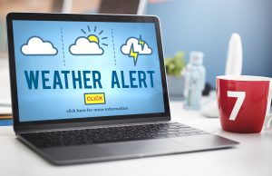 Laptop on a desk displaying a "Weather Alert" screen with cloud, sun, and lightning icons and a yellow "Click" button, next to a red coffee mug marked with the number 7.
