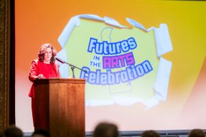 A woman in a red outfit speaks at a wooden podium with a microphone on a theater stage. Behind her, a large projected graphic reads “Futures in the Arts Celebration” with a Delaware outline on a yellow-to-orange background.