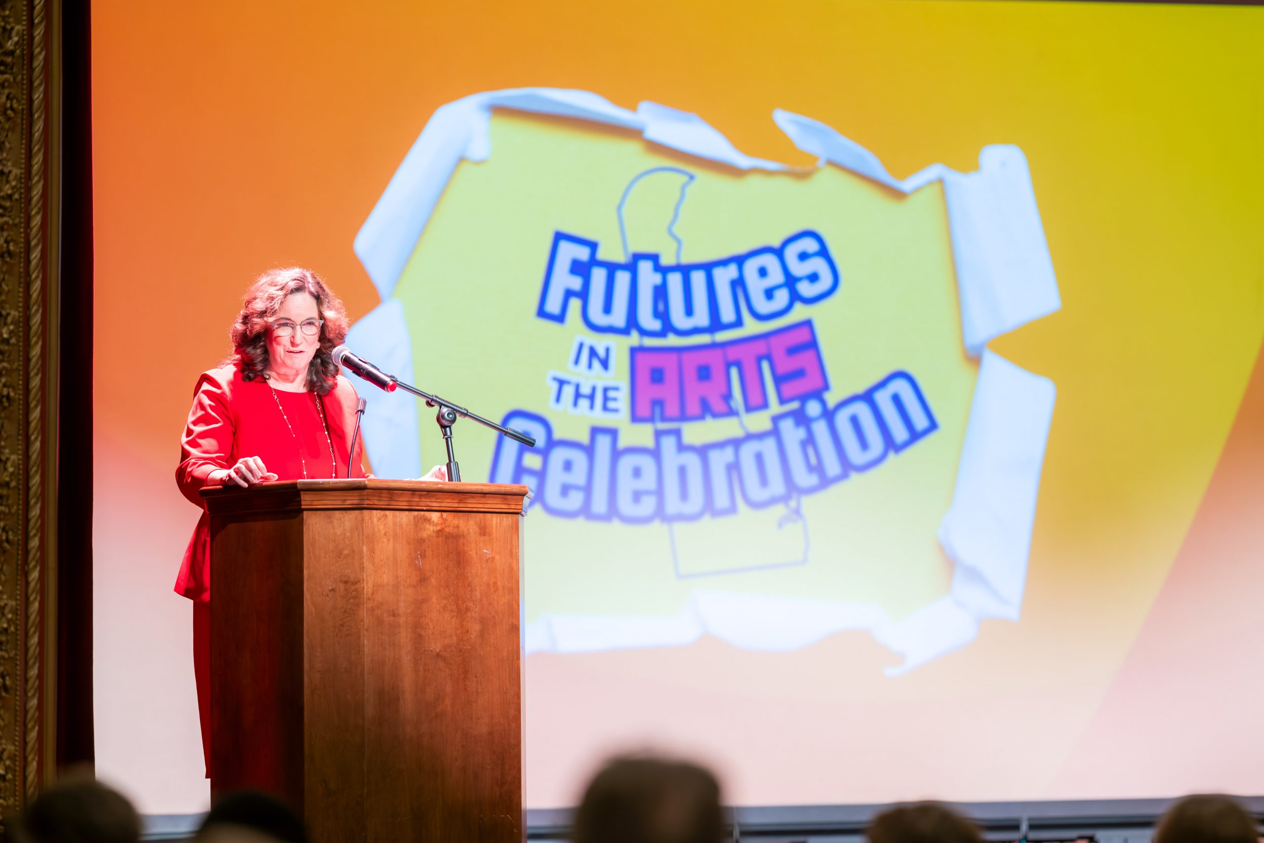 A woman in a red outfit speaks at a wooden podium with a microphone on a theater stage. Behind her, a large projected graphic reads “Futures in the Arts Celebration” with a Delaware outline on a yellow-to-orange background.