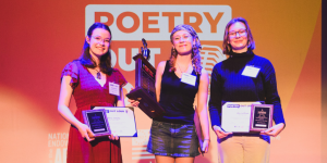 Three students stand on a stage in front of a large “Poetry Out Loud” backdrop, smiling and holding certificates and award plaques; the student in the center holds a large trophy/award while the two on either side hold framed plaques and certificates under colorful stage lighting.