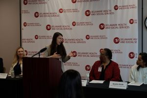 Woman stands behind a brown podium in front of a white and red back drop giving a presentation. One woman is behind a table on the left. Two other women are behind another table to the right.