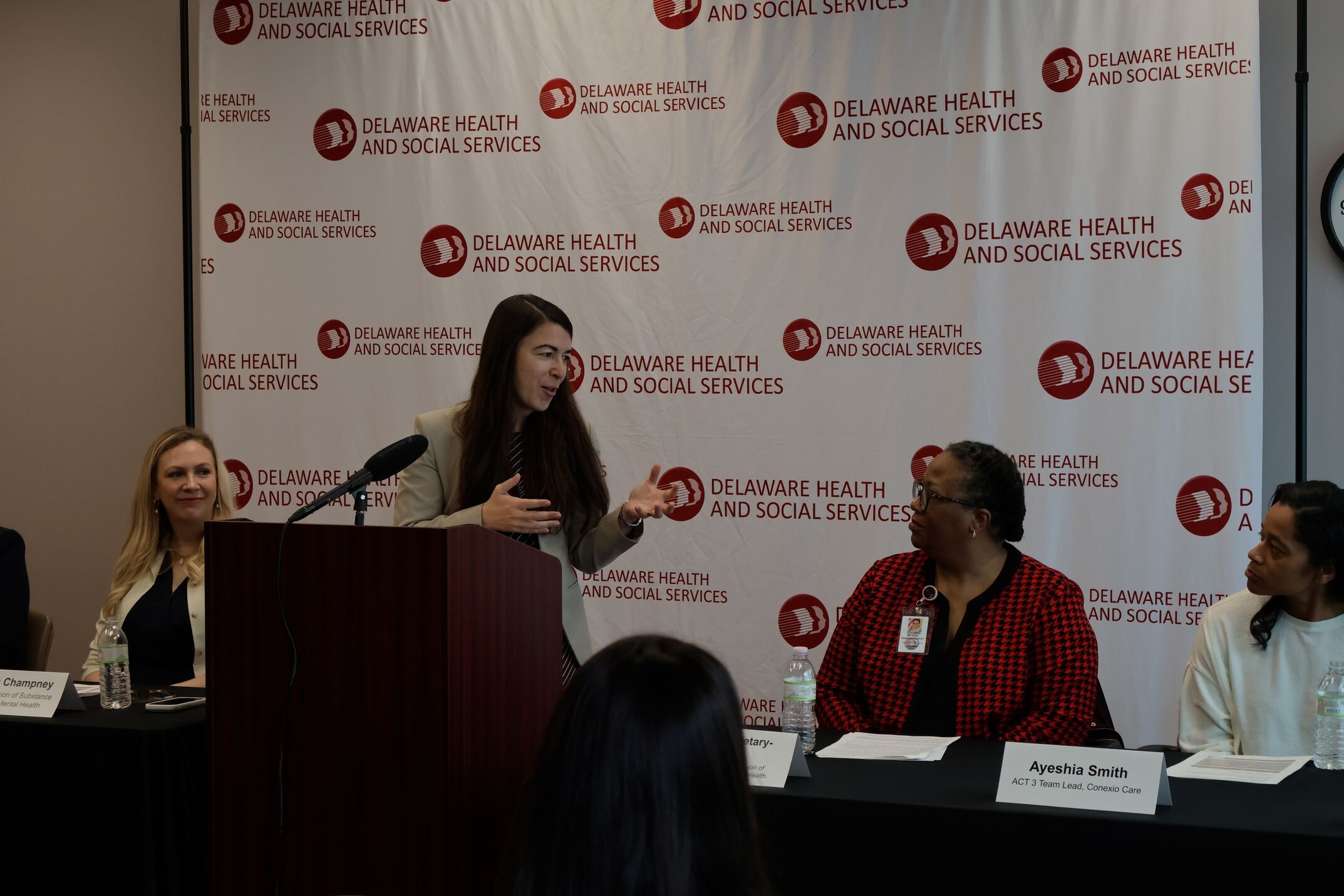 Woman stands behind a brown podium in front of a white and red back drop giving a presentation. One woman is behind a table on the left. Two other women are behind another table to the right.