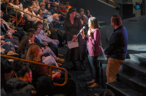 A woman stands at the front of an auditorium speaking to a large audience of teenage students.