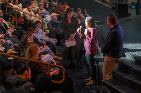 A woman stands at the front of an auditorium speaking to a large audience of teenage students.