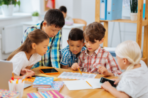 Five children sit and stand in a huddle around a table reading the same document.