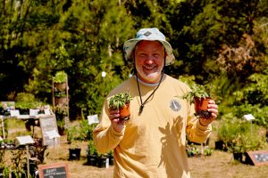 Man in a bucket hat and long sleeve yellow shirt smiles at the camera holding two small plants, in front of many plants for sale in the background.