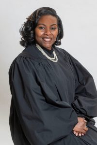 A professional headshot of a woman wearing a black judicial robe and a pearl necklace, smiling warmly against a neutral gray background. Her hair is styled in soft curls, and her hands are gently folded in front of her.