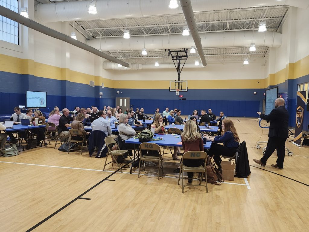 A large group seated at round tables in a gymnasium attending a presentation by a speaker.