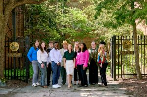 A group of ten professionals stand in front of a wrought iron gate labeled "DNREC Courtyard Visitor Entrance." They are outdoors, surrounded by greenery and trees, with a brick building in the background.