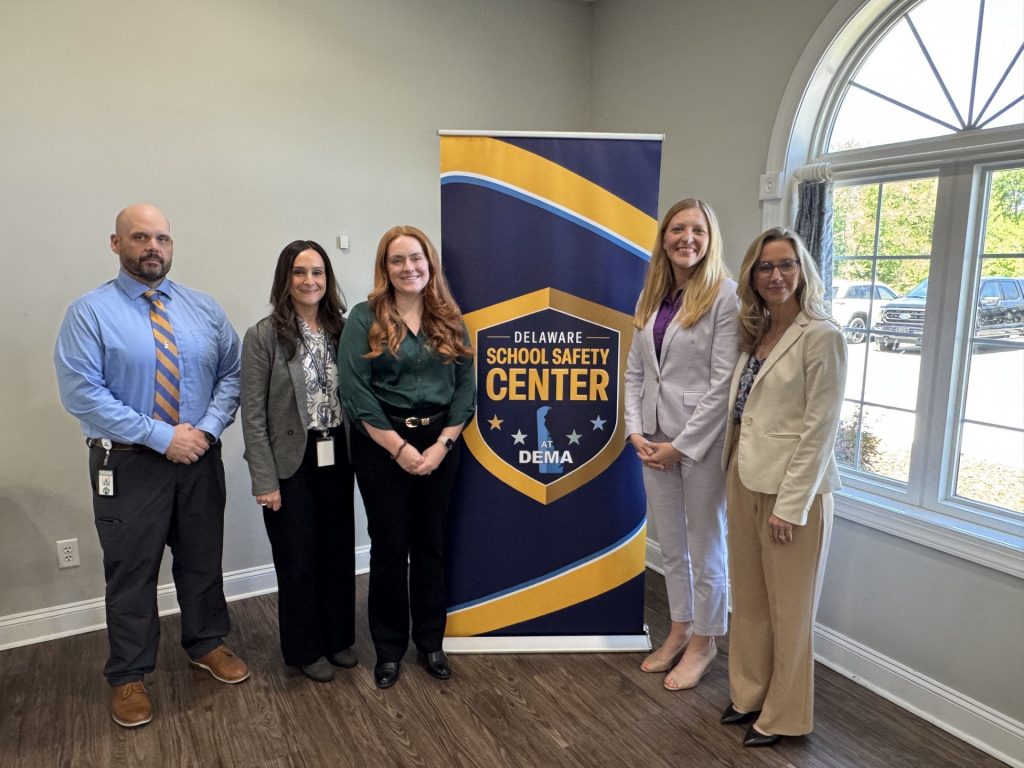 Members of the Delaware School Safety Center and Delaware Department of Education gathered around a Delaware School Safety Center display in a well-lit room with arched window.