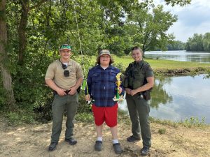Gabriel Alfaro, winner of the 2026 Youth Fishing Tournament in Delaware, stands between a male Delaware Natural Resources Police officer on the left, and a female officer on the right, holding his winning trophy and fishing pole by Lums Pond.