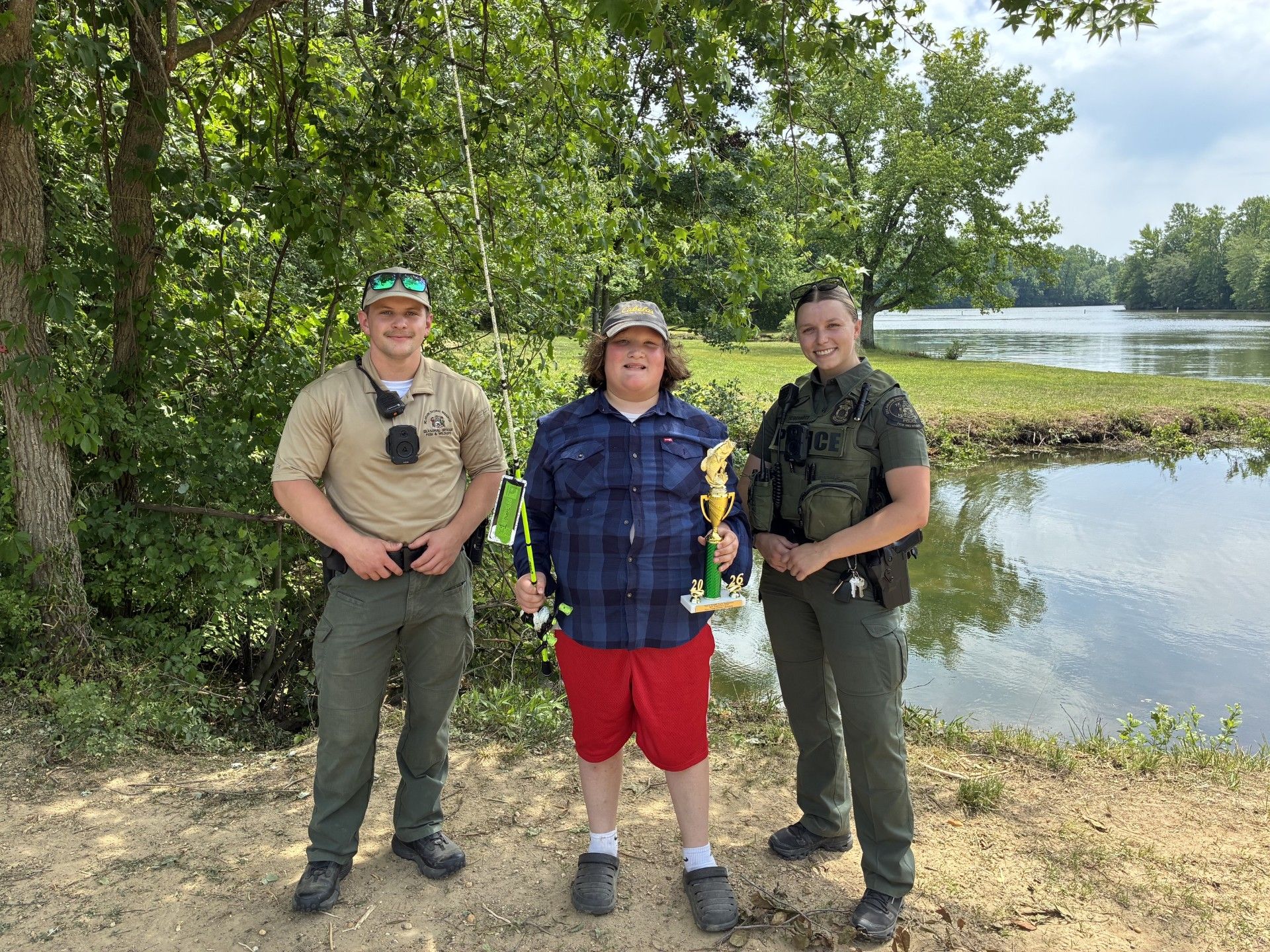 Gabriel Alfaro, winner of the 2026 Youth Fishing Tournament in Delaware, stands between a male Delaware Natural Resources Police officer on the left, and a female officer on the right, holding his winning trophy and fishing pole by Lums Pond.