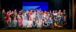 Three rows of about 25 students stand on a stage, smiling at the camera.