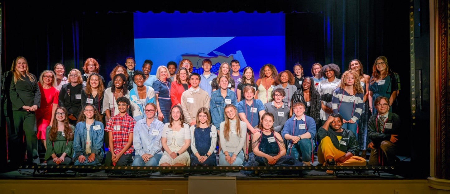 Three rows of about 25 students stand on a stage, smiling at the camera.