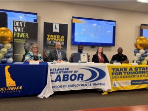 Adults sit at tables during a career fair. The tables have signs on them that say, "Join Delaware Schools. Department of Labor. Take a Stand with a Teaching Career in Delaware."