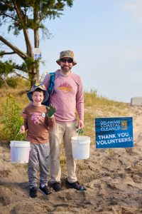 Two volunteers, a father and their child, stand next to the Coastal Cleanup "Thank you, Volunteers" sign, with beach in the background.