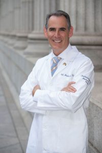 Dr. Neil Hockstein, in a white lab coat and tie stands with his arms crossed in front of a stone-columned building.