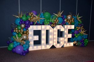 Large, lighted, block letters that spell EDGE are surrounded by balloon art. There are cyan blue, gold, purple and gold round balloons, with spikey gold balloons interspersed.