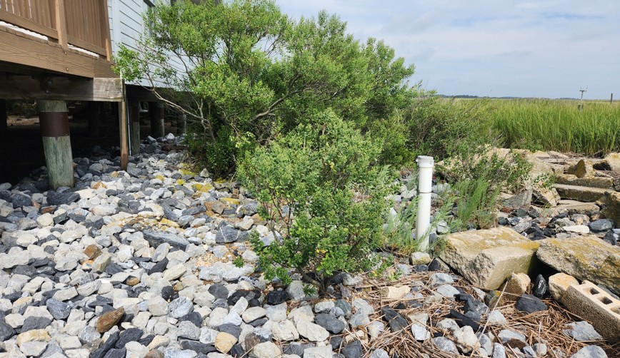 A white private well pipe stands beside a property amid rocks and vegetation near a coastal marsh.