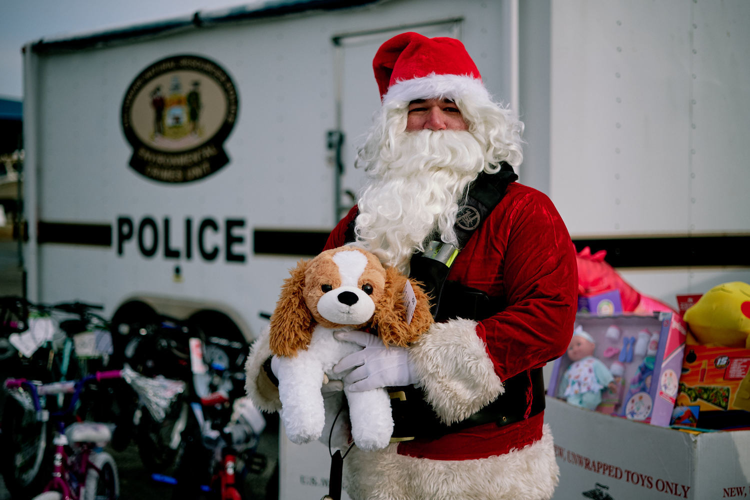 Santa Claus holds a stuffed dog plush in front of the boxes of Toys for Tots donations collected in 2025 and the Delaware Natural Resources Police trailer.