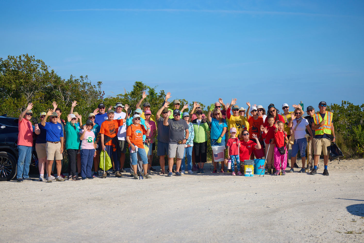 Group photo of about 36 people waving toward the camera at the 39th Coastal Cleanup. The stand with the beach dunes in the background